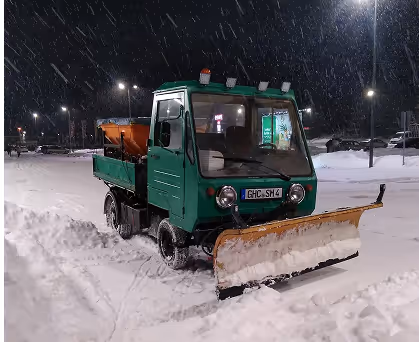 Kleiner grüner Schneepflug fährt bei Nacht im Schneefall auf einer schneebedeckten Straße.